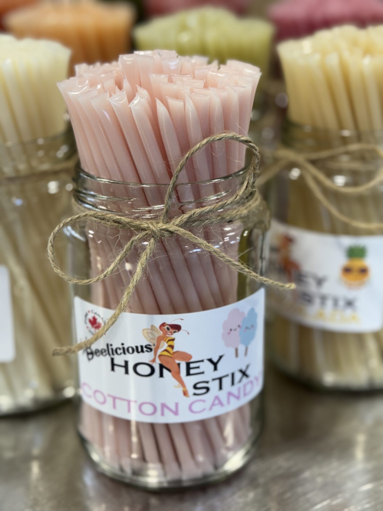 A jar of pink honey sticks tied with twine labeled 'Honey Stix Cotton Candy.'